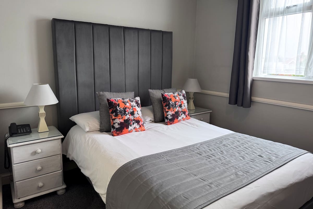 Una cama bien hecha con colcha gris, dos almohadas de estampado floral, una lámpara de noche y una mesita de noche con un teléfono.