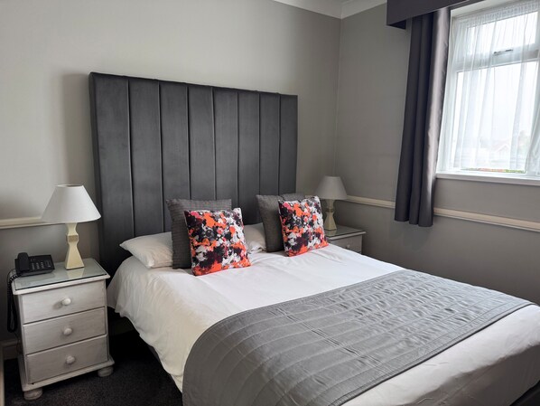 A neatly made bed with a grey quilt, two pillows with a floral pattern, a bedside lamp, and a nightstand with a phone. - Camelia Hotel (Southend-on-Sea)