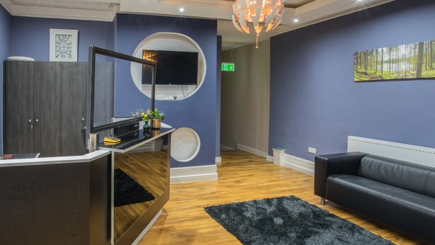 A reception area with a wooden reception desk, a black sofa, a black rug, a round mirror, and a flat-screen TV mounted on a wall. - High Bank Hotel (Leeds)