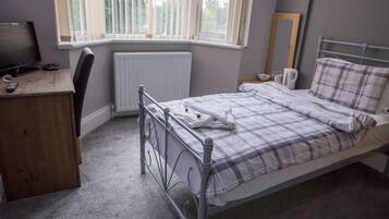 A bedroom with a bed, a desk, a chair, a radiator, a window with blinds, a mirror, and a bedside table with a kettle and glasses.