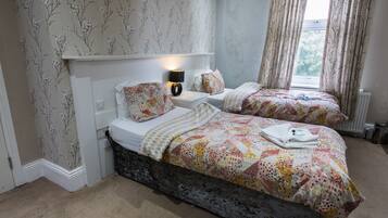 A bedroom with two beds, floral wallpaper, a nightstand with a lamp, and a window with curtains.