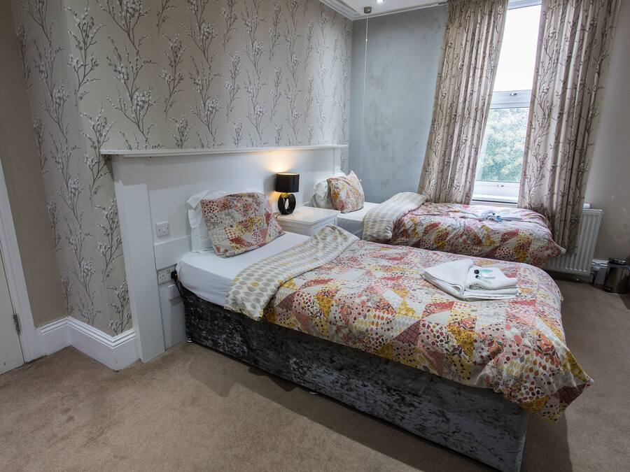 A bedroom with two beds, floral wallpaper, a nightstand with a lamp, and a window with curtains.