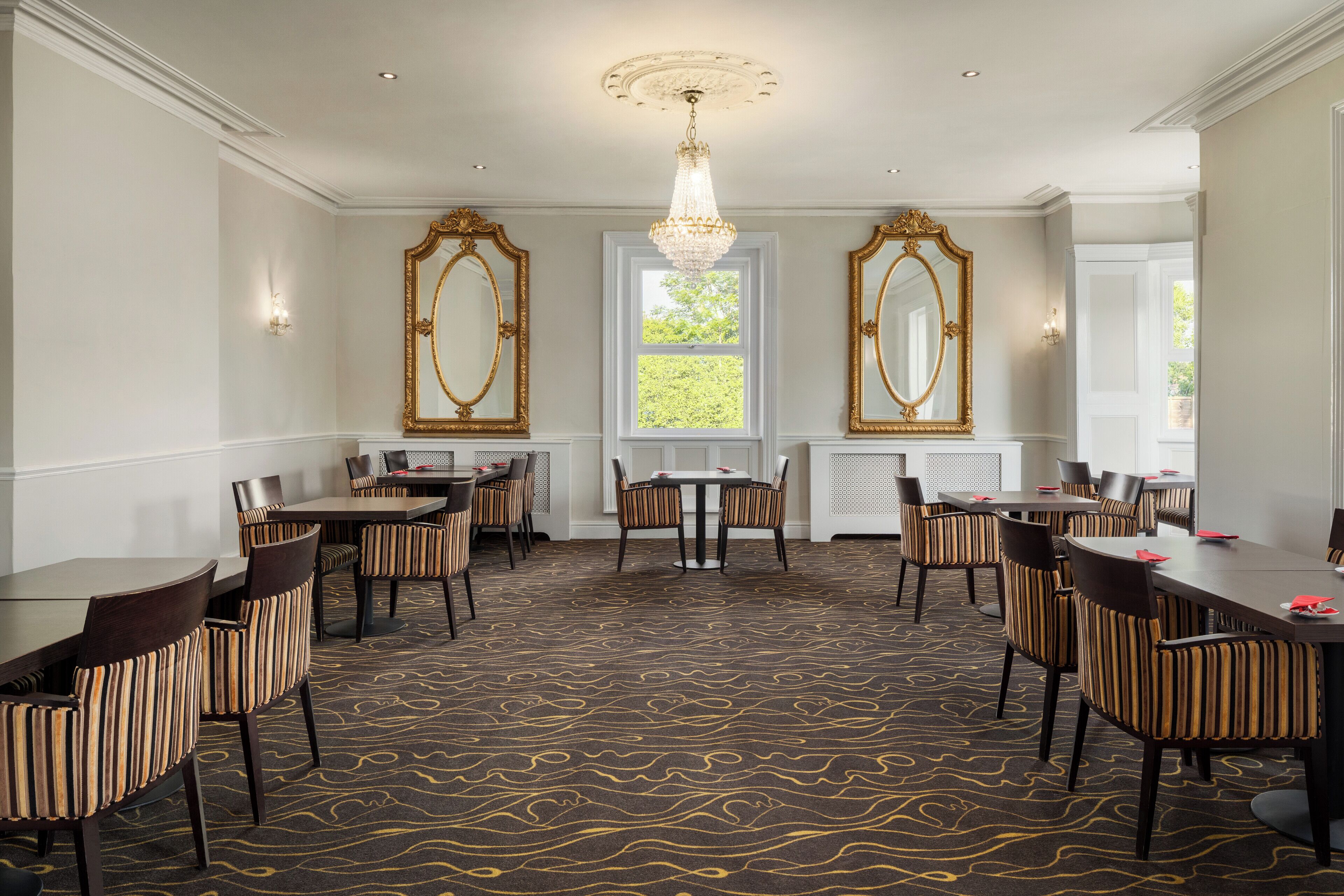 A hotel dining area with multiple tables and chairs, ornate mirrors, and a chandelier.