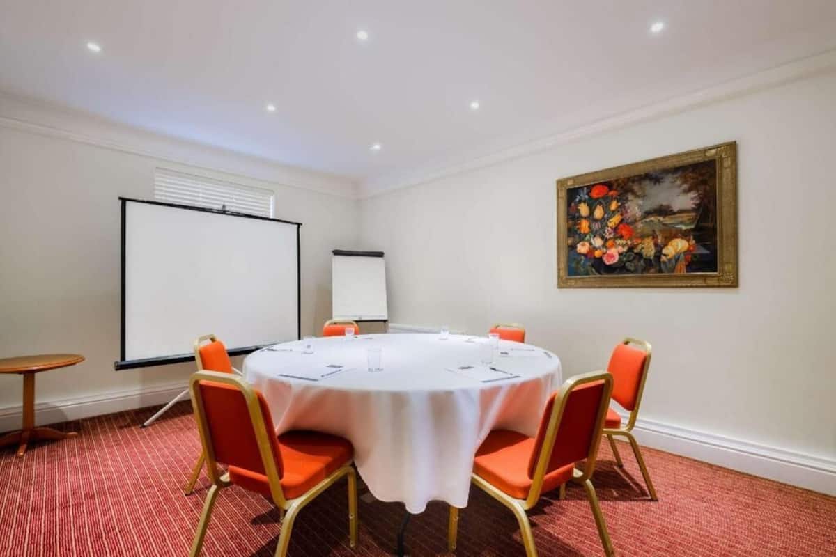A conference room with a round table, four chairs, a projector screen, and a painting on the wall.