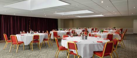 Une salle de conférence dotée de plusieurs tables rondes, chacune recouverte d’une nappe blanche et entourée de chaises rouges.