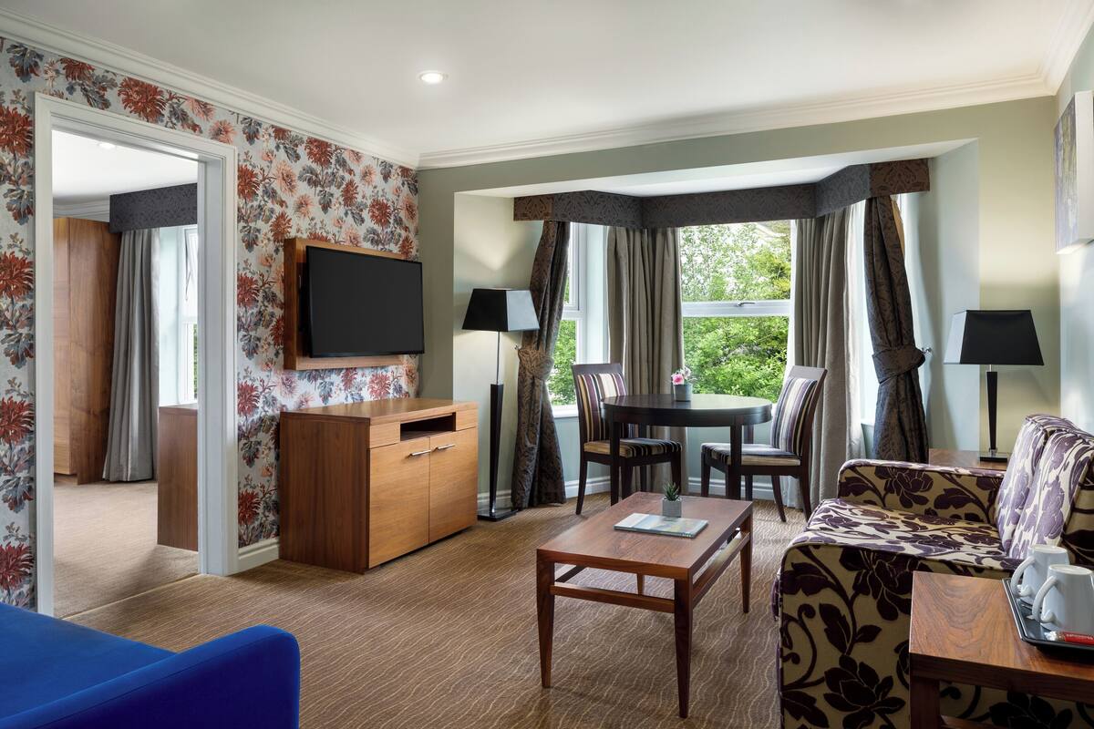 A hotel room with a wooden desk, a sofa, a dining table, and a TV mounted on the wall.