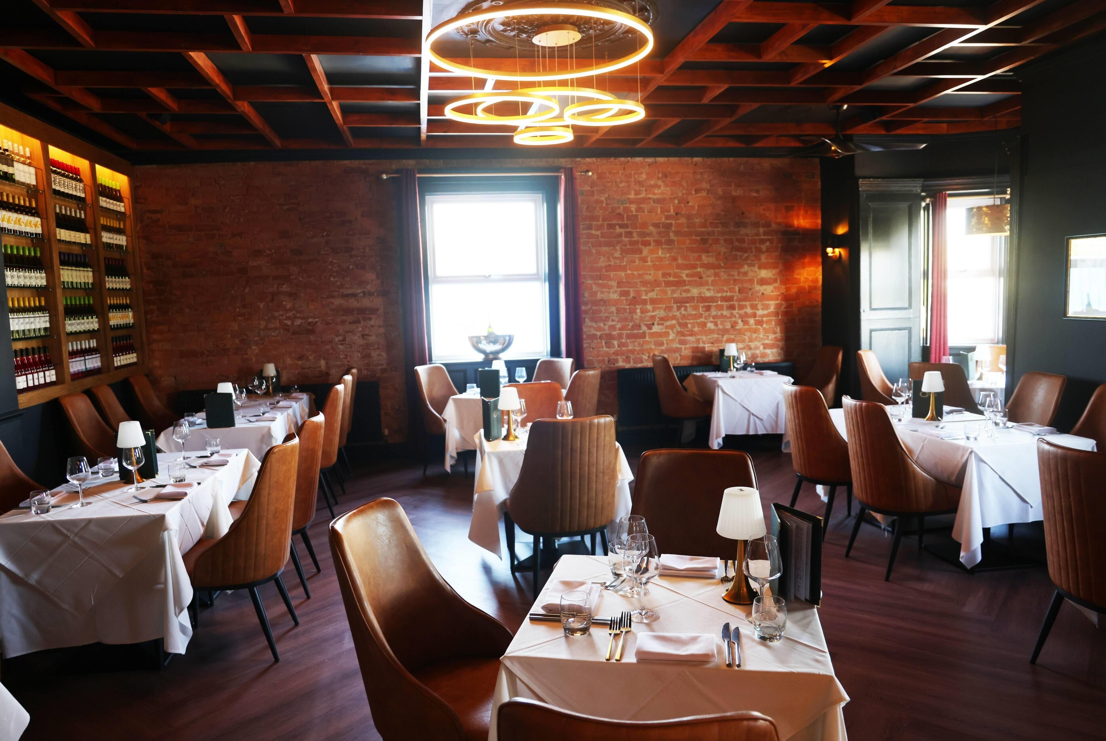 A restaurant interior with brick walls, wooden beams, and a modern chandelier. Tables are set with white tablecloths and brown chairs.