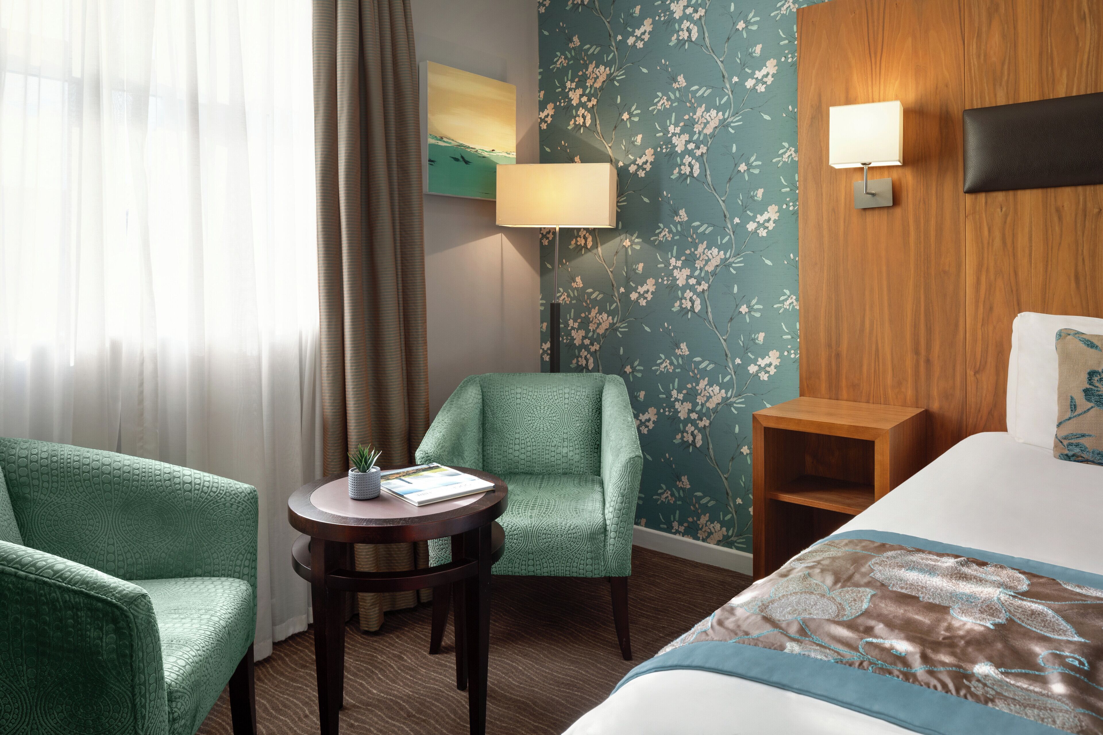 A hotel room with a bed, two armchairs, a small table with a lamp, and a wall with floral wallpaper.