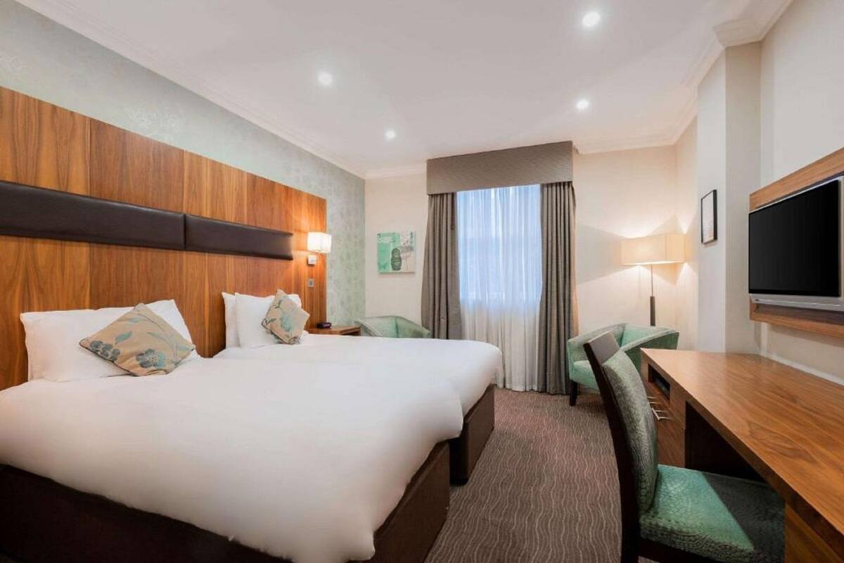 A hotel room with a large bed, a desk, two chairs, a TV, and a window with curtains.
