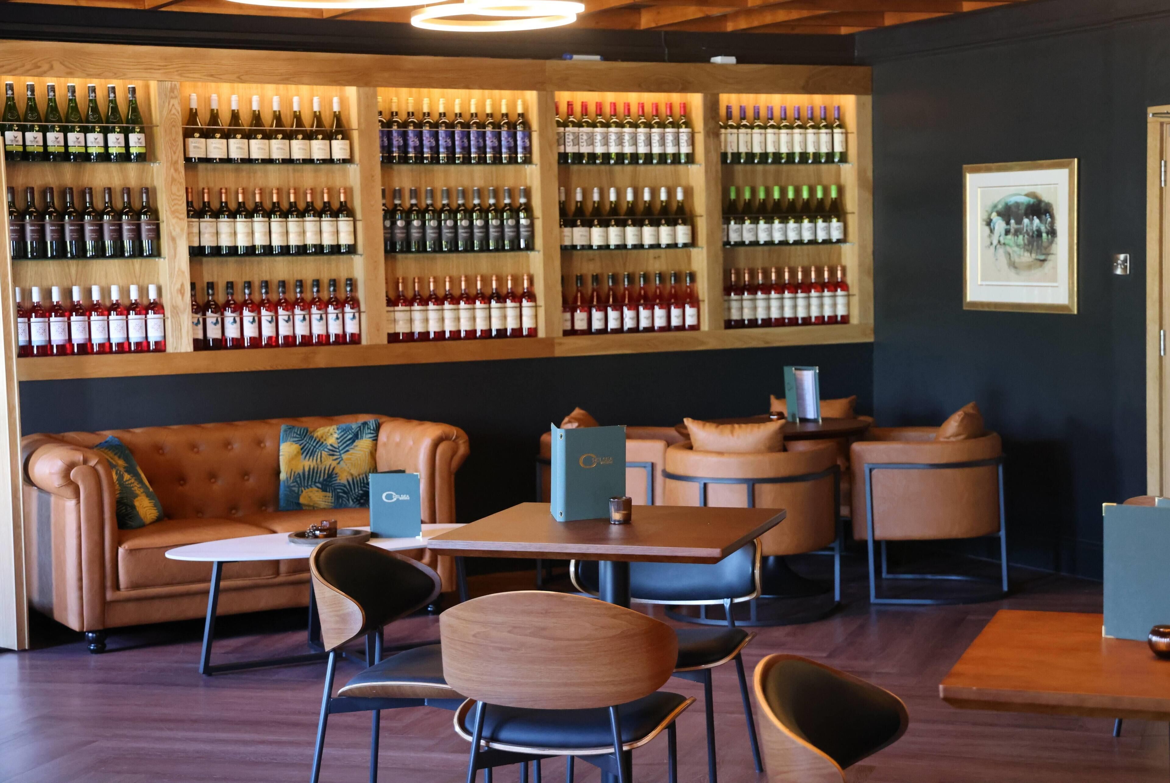 Zona de bar con varias opciones de asiento, incluyendo un sofá y mesas, y una pared llena de botellas de vino.