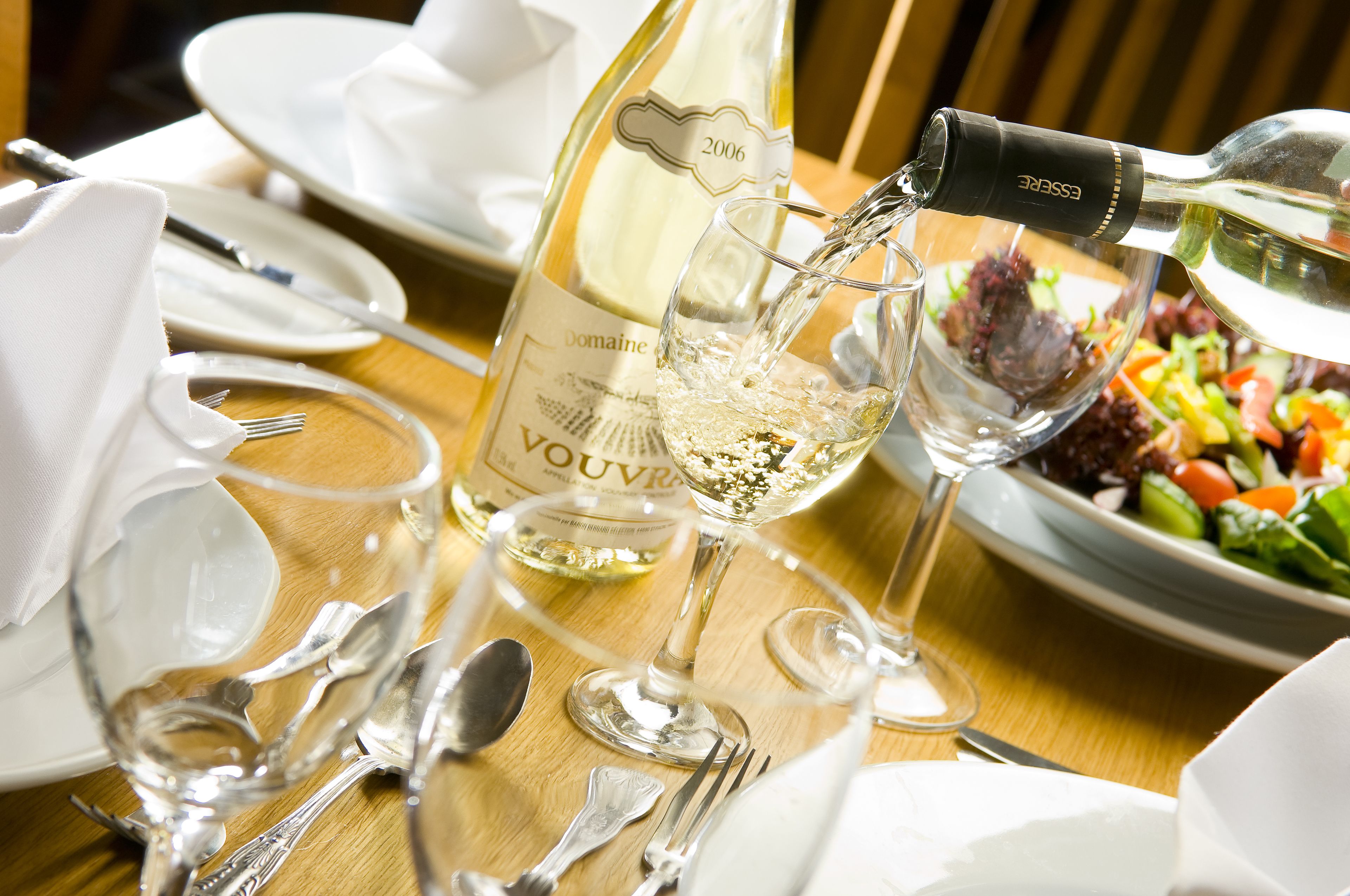 A dining table set with white tableware, a bottle of Domaine Vouvray wine, and a glass of white wine being poured.