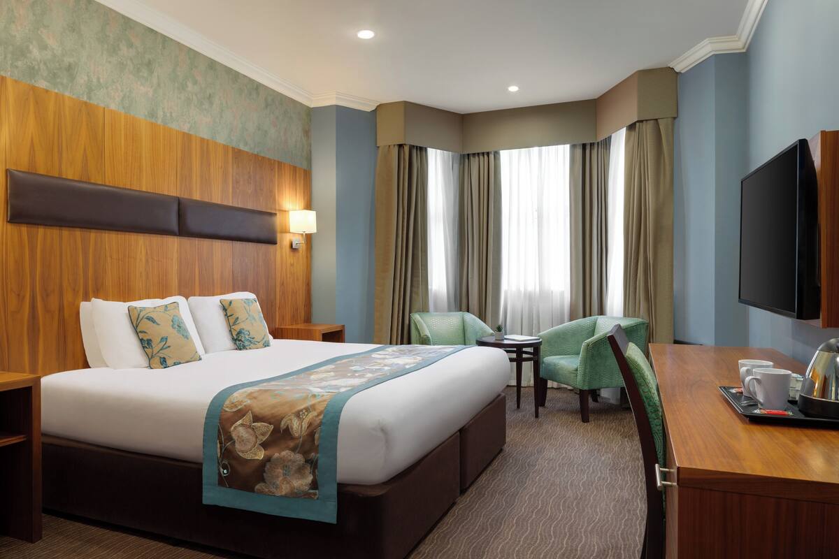 A hotel room with a large bed, a desk with a chair, a television, and a window with curtains.