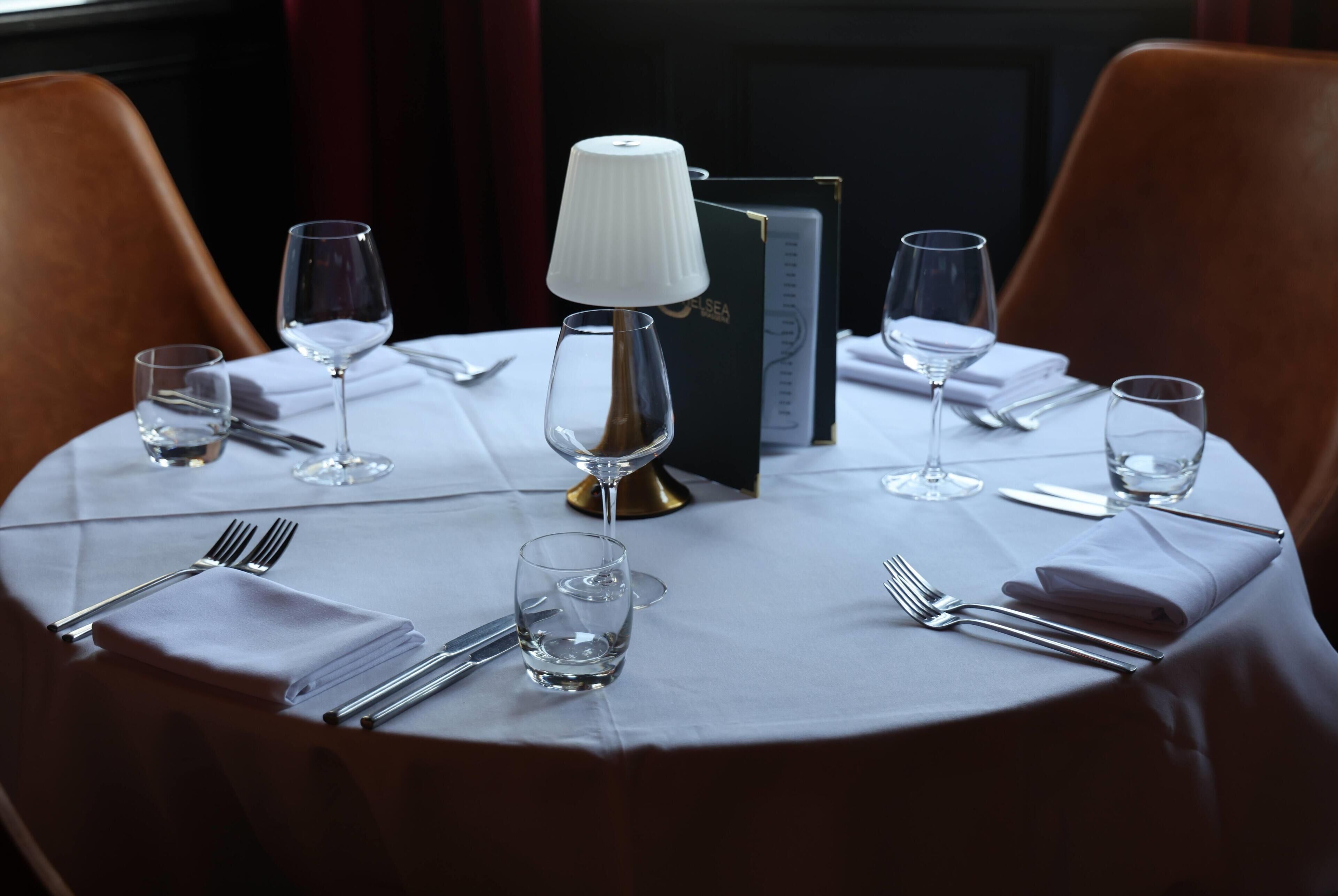 A round dining table with a white tablecloth, set with wine glasses, cutlery, and a lamp.