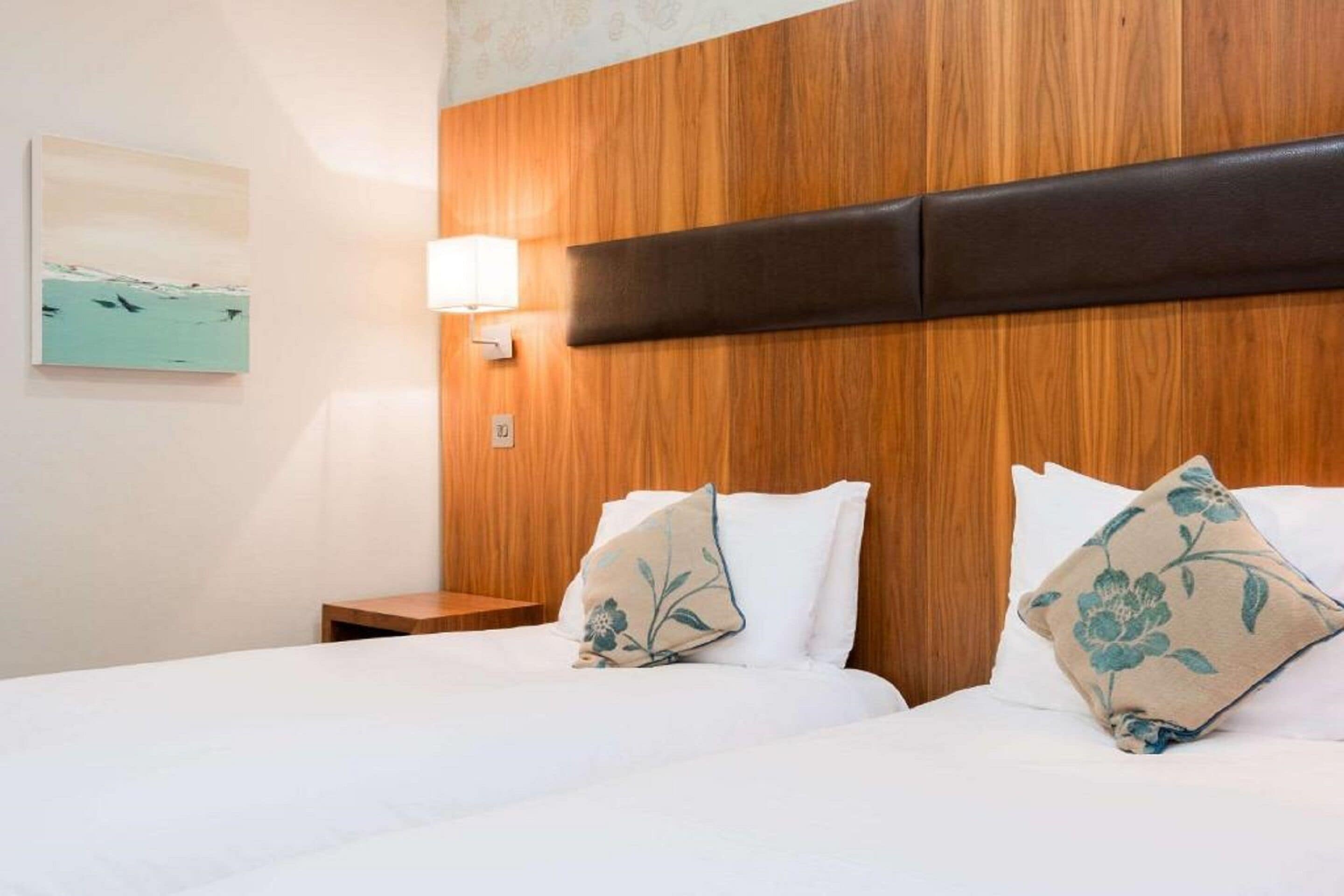 Ein Hotelzimmer mit einem hölzernen Kopfteil, zwei Betten mit weißen Laken, einem Nachttisch, einer Wandleuchte und einem Kissen mit Blumenmuster.