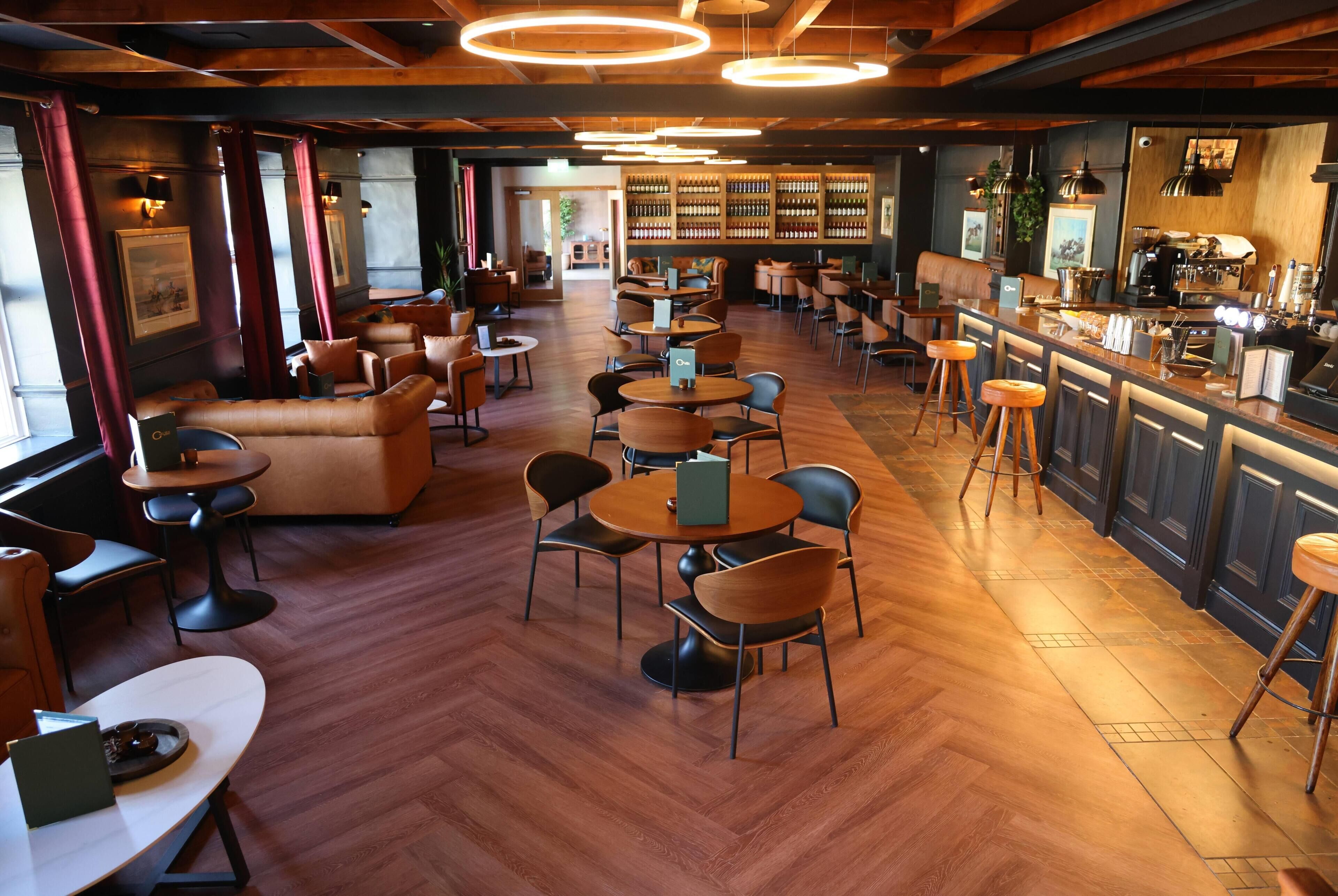 Eine Barbereich mit Holzboden, runden Tischen, Ledersitzmöbeln und einer Bar mit einer Kaffeemaschine.