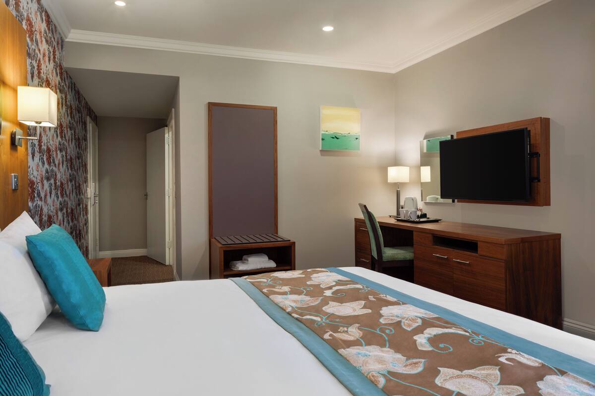 A hotel room with a bed, a desk, a chair, a television, and a painting on the wall.