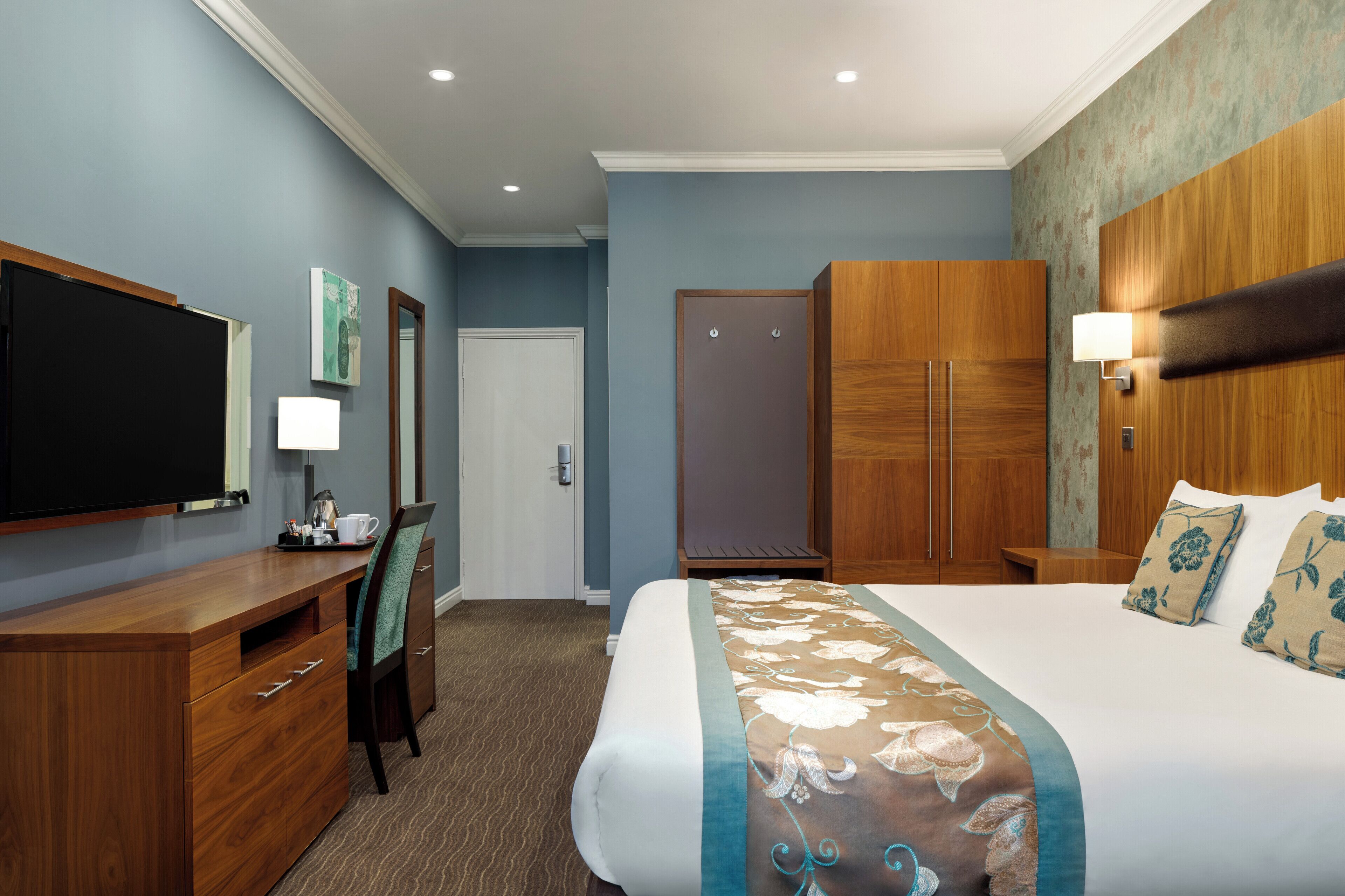 A hotel room with a large bed, a wooden desk, a television, a wardrobe, and a bathroom door.