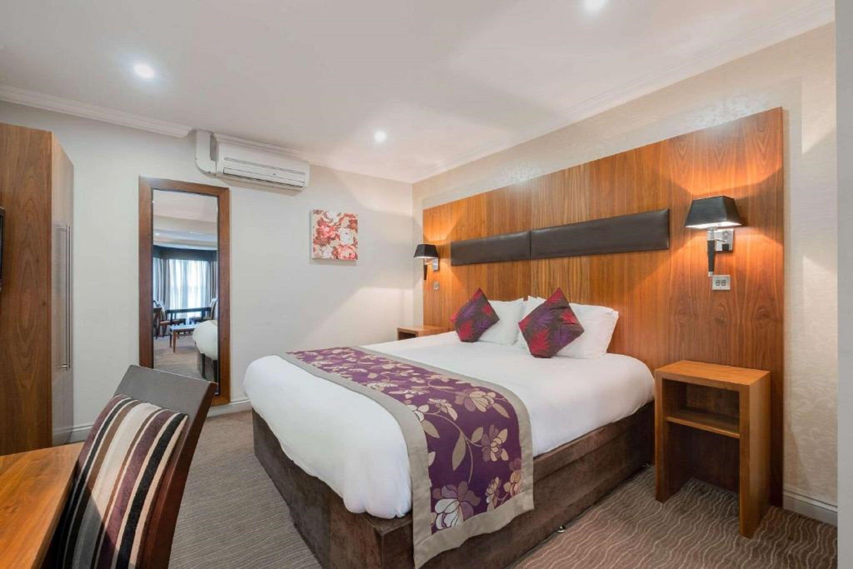 Habitación de hotel con cabecera de madera, una cama grande con colcha floral púrpura, dos mesitas de noche con lámparas y vista al área de comedor a través de una puerta abierta.