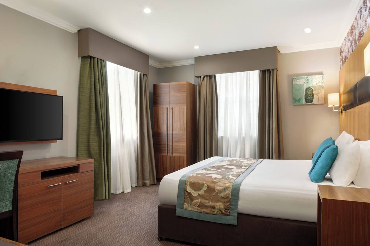 A hotel room with a bed, a TV, a wooden wardrobe, and a painting on the wall.