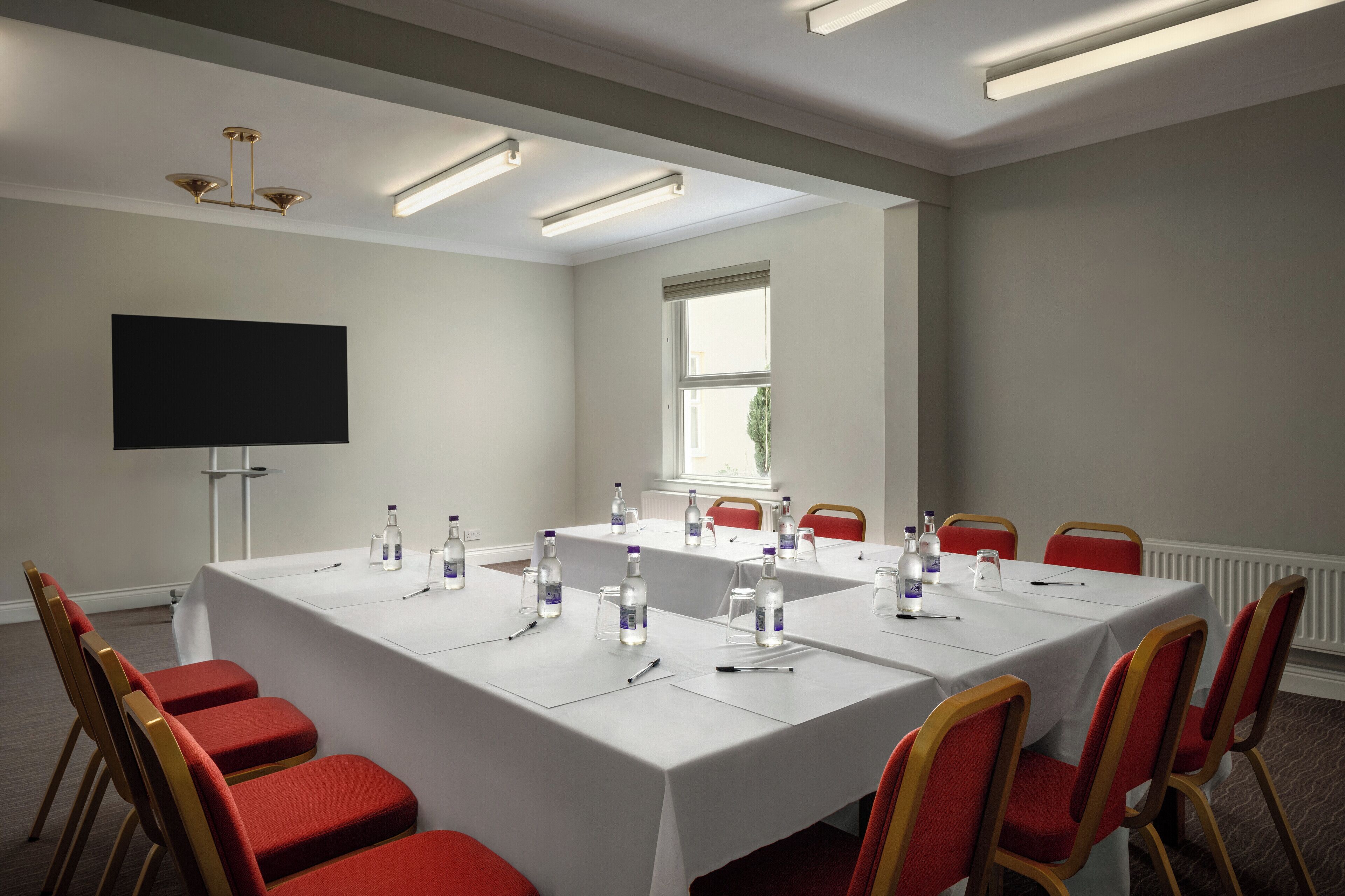 A conference room with a long table, red chairs, water bottles, and a flat-screen TV.