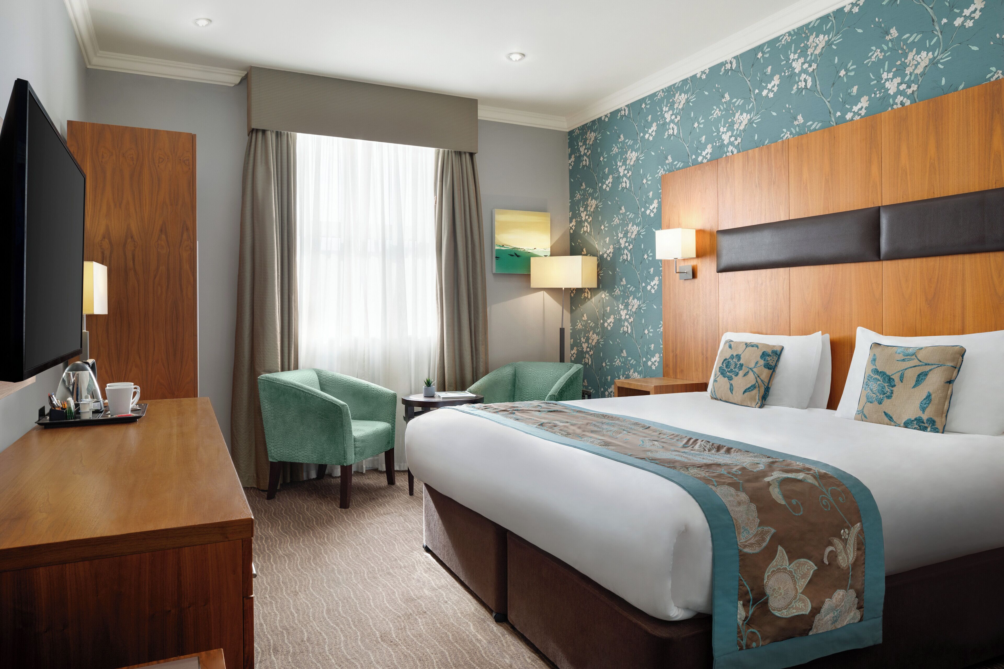 Habitación de hotel con una cama grande, un escritorio con televisor, una silla y una mesita con lámpara.