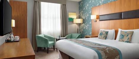 Habitación de hotel con una cama grande, un escritorio con televisor, una silla y una mesita con lámpara.