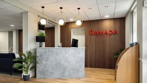 Ein Empfangsbereich mit einer Rezeption, einem blauen Sofa und einer Holzwand mit dem Schriftzug 'RAMADA' in roten Buchstaben.
