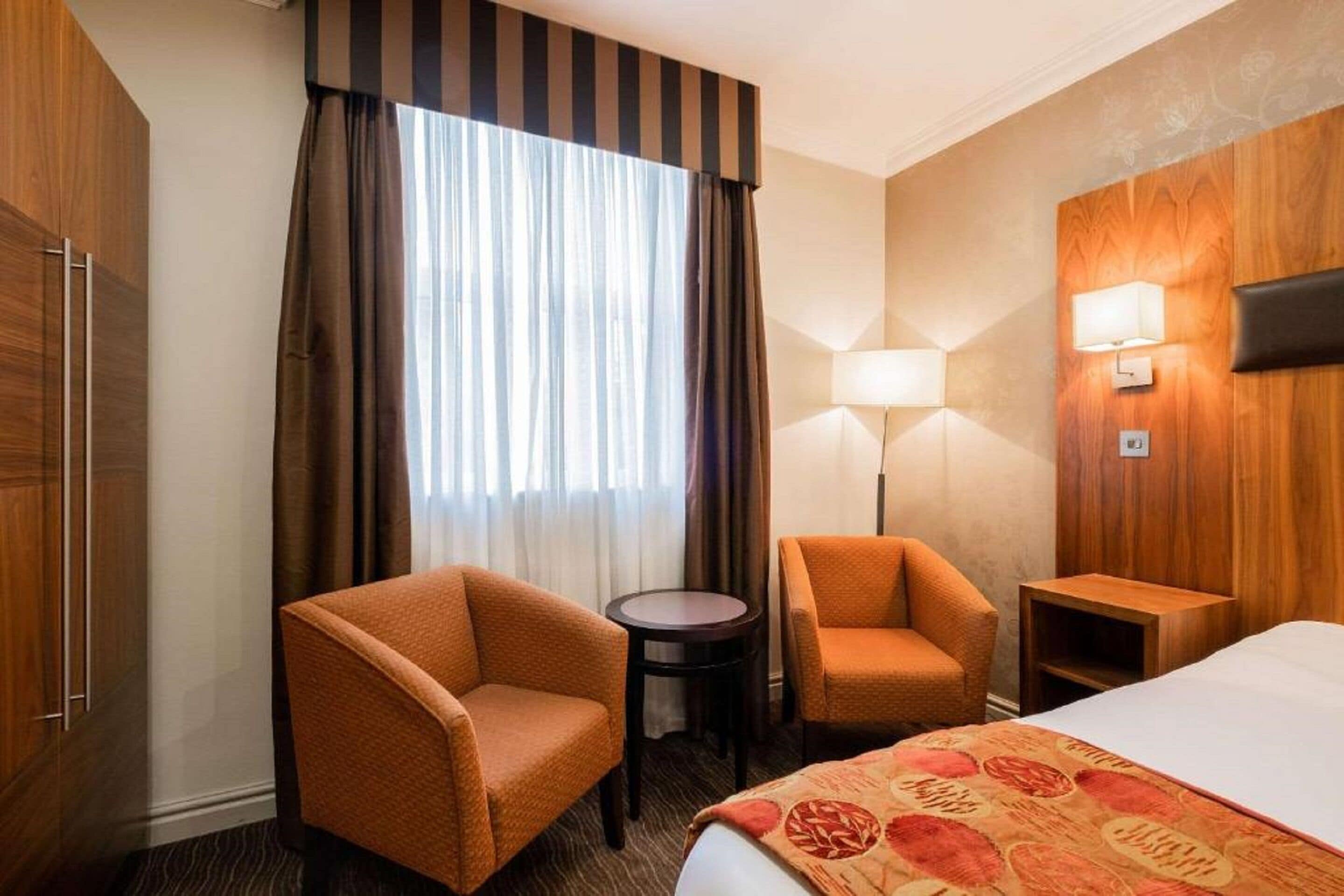 A hotel room with a bed, two armchairs, a small table, a lamp, and a view of the outdoors through a window with curtains.
