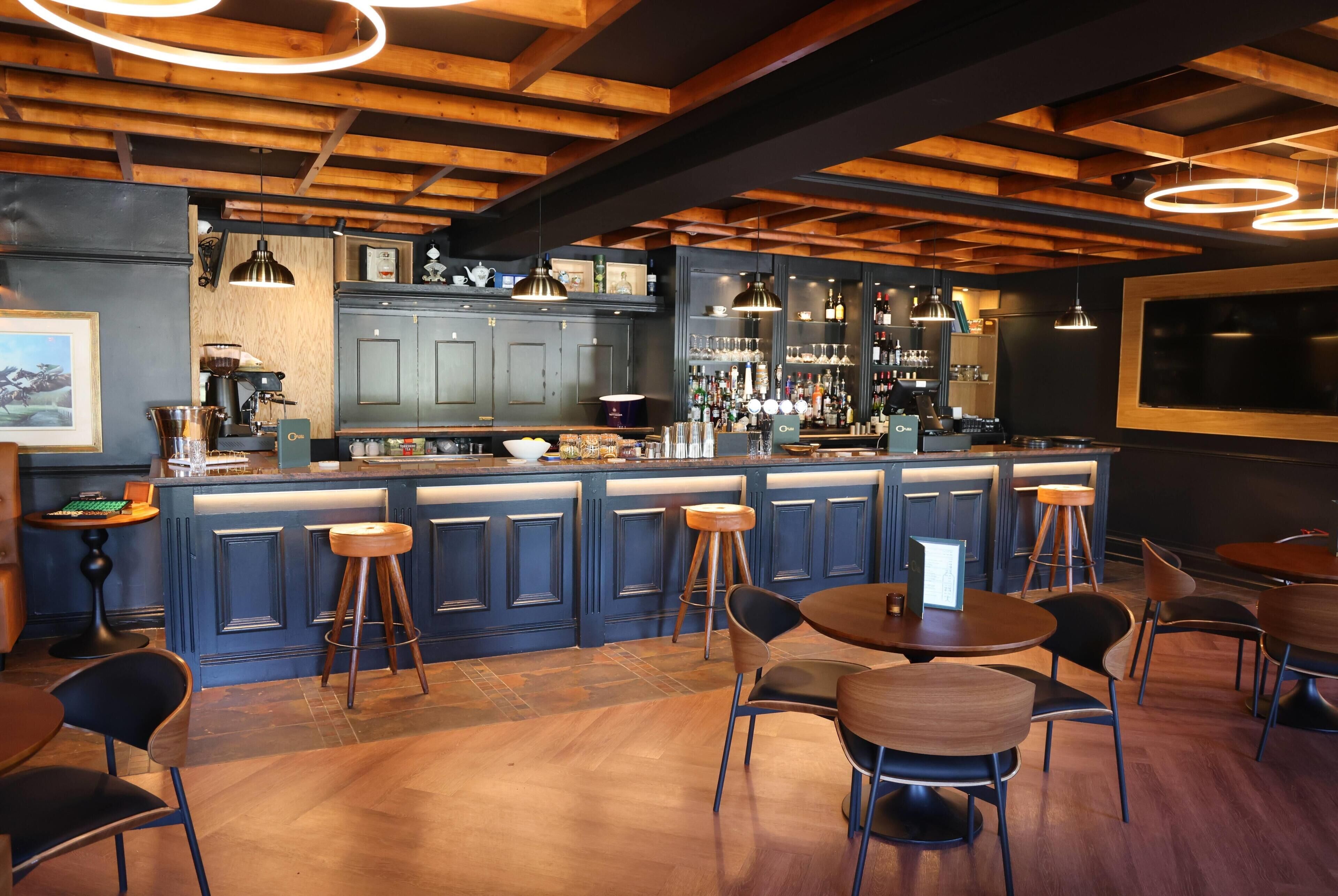 Zona de bar con vigas de madera, taburetes y una barra con diferentes botellas y vasos.