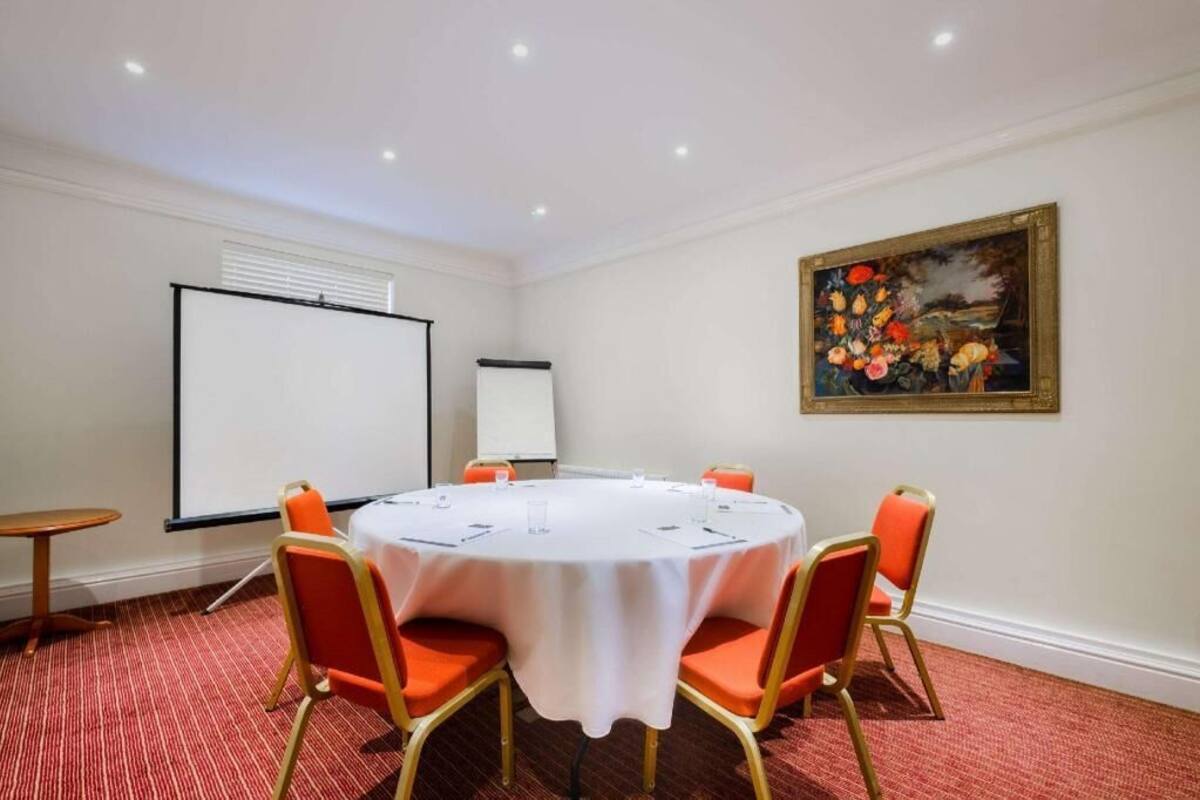 A conference room with a round table, four chairs, a projector screen, and a painting on the wall.
