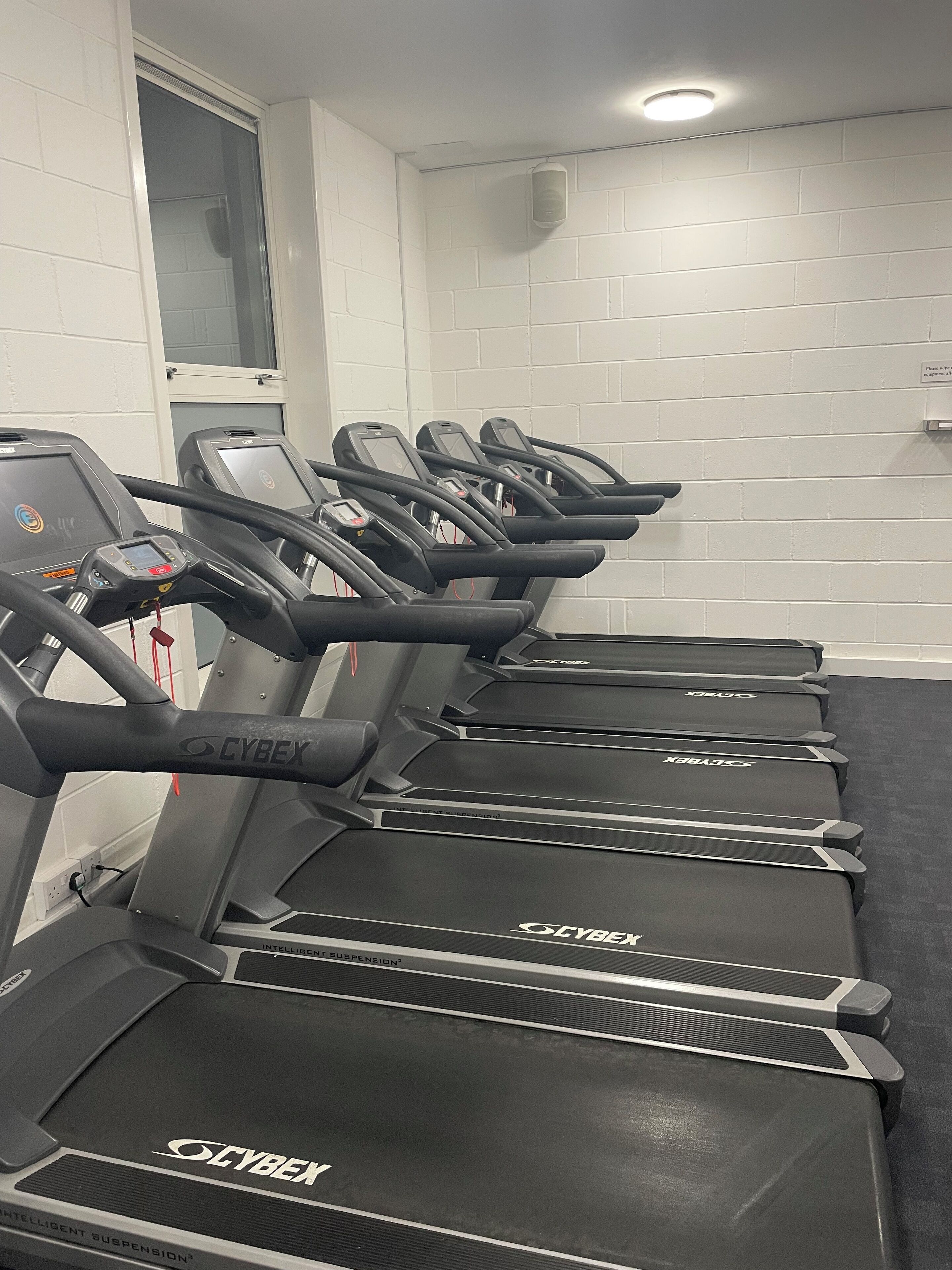 A row of Cybex treadmills in a gym with white brick walls and a window.
