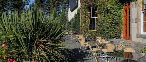 An outdoor patio with wicker chairs and tables, surrounded by greenery and a building with a stone facade.
