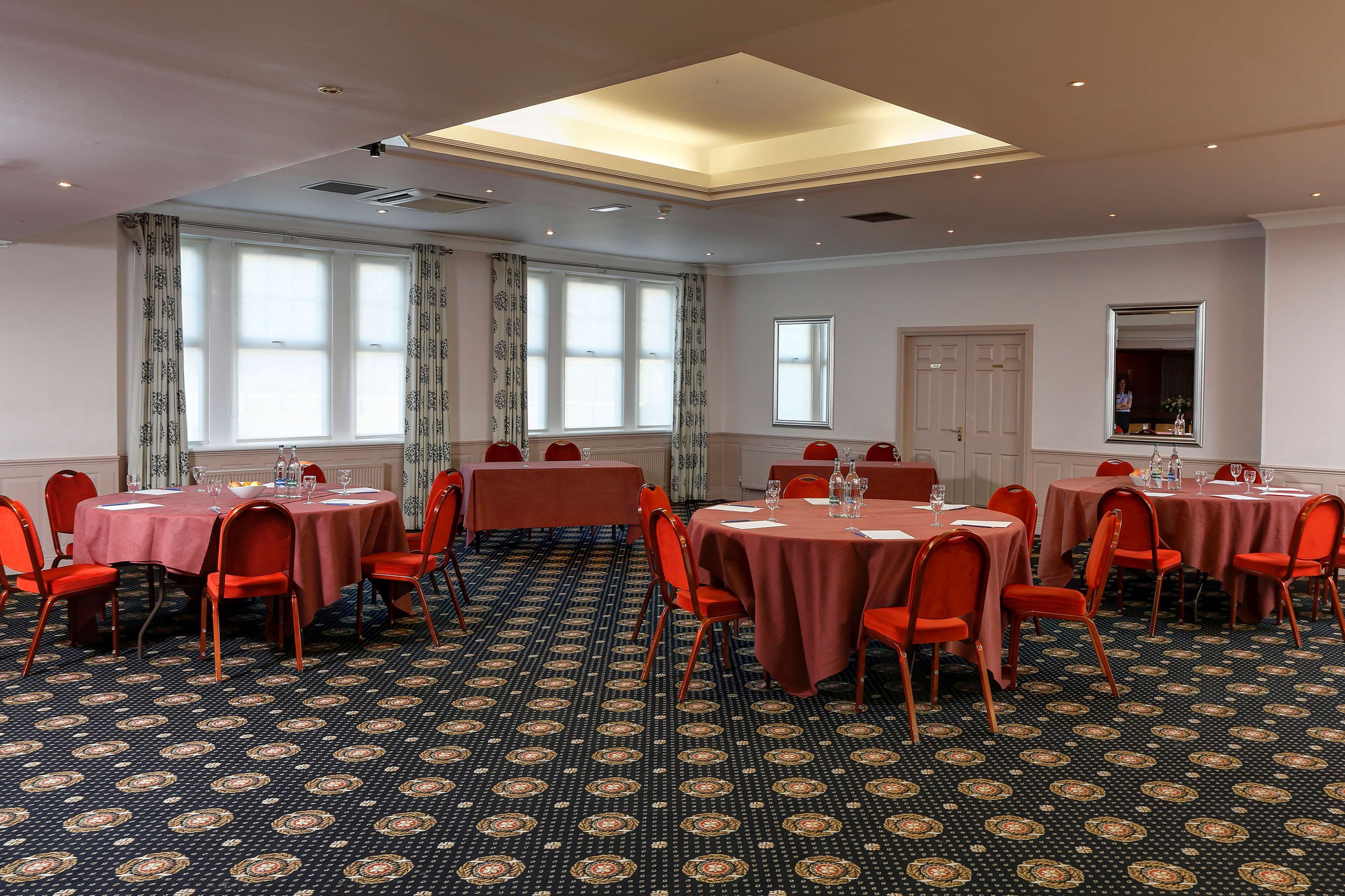 Un salón de conferencias con varias mesas y sillas, amplias ventanas con cortinas y una alfombra estampada.