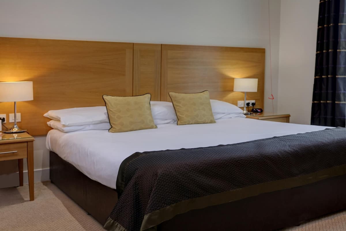 A hotel room with a large bed, two bedside lamps, a wooden headboard, and a window with curtains.