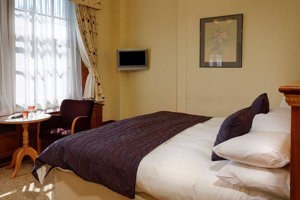 A hotel room with a bed, a television, a chair, a small table, and a window with curtains.