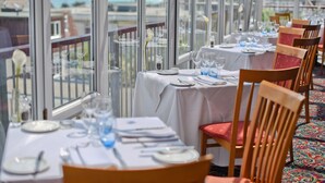 Un restaurant amb taules preparades per dinar, grans finestres que donen a una vista del mar i una catifa colorida.