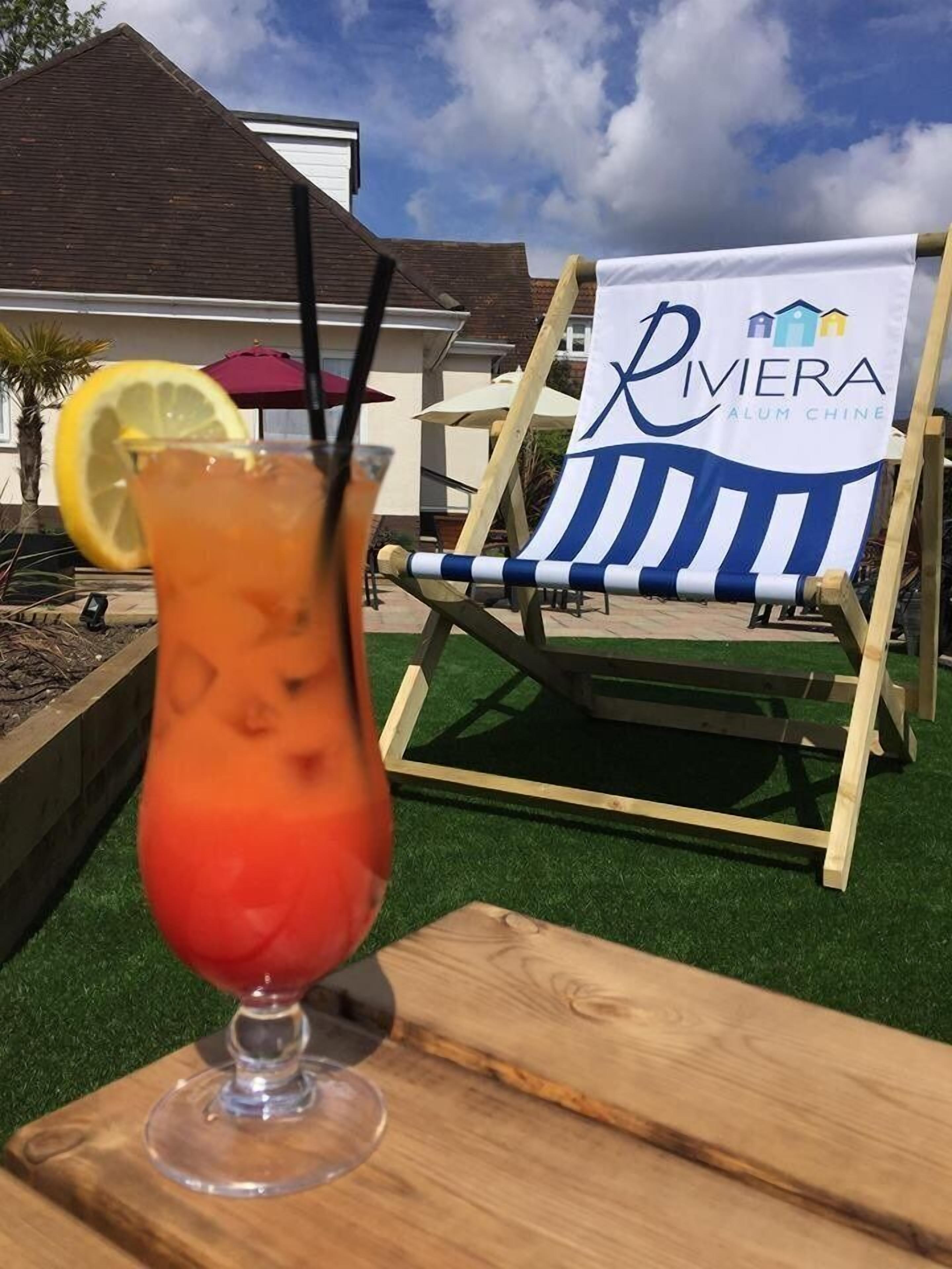 A wooden table with a cocktail glass and a striped beach chair in the background. A banner with the text "Riviera Alum Chine" is visible.