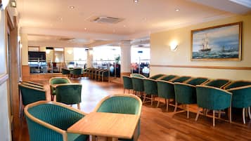 A spacious dining area with green upholstered chairs and wooden tables, large windows, and a painting of a ship on the wall.