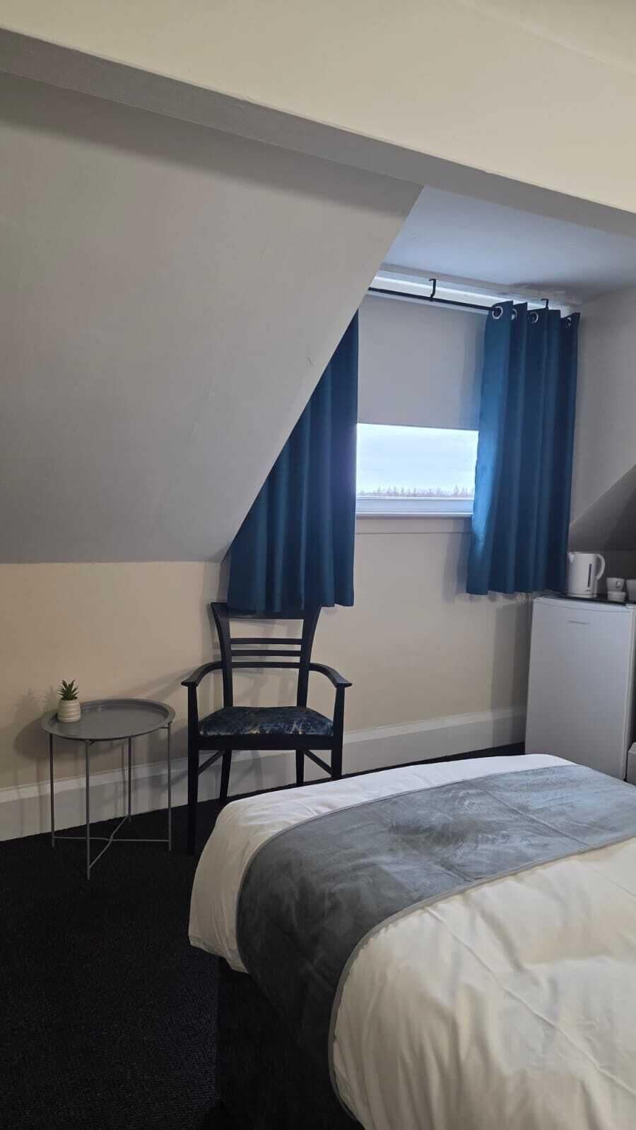 A hotel room with a bed, a chair, a small table, and a window with curtains.