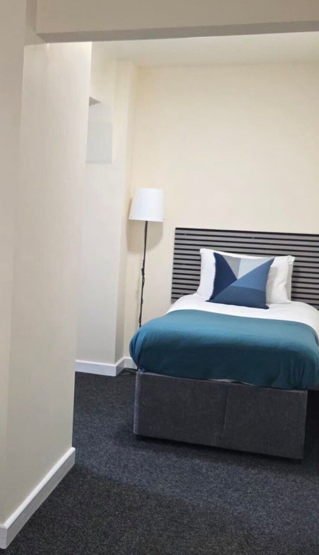 A hotel room with a bed, a bedside lamp, and a carpeted floor.