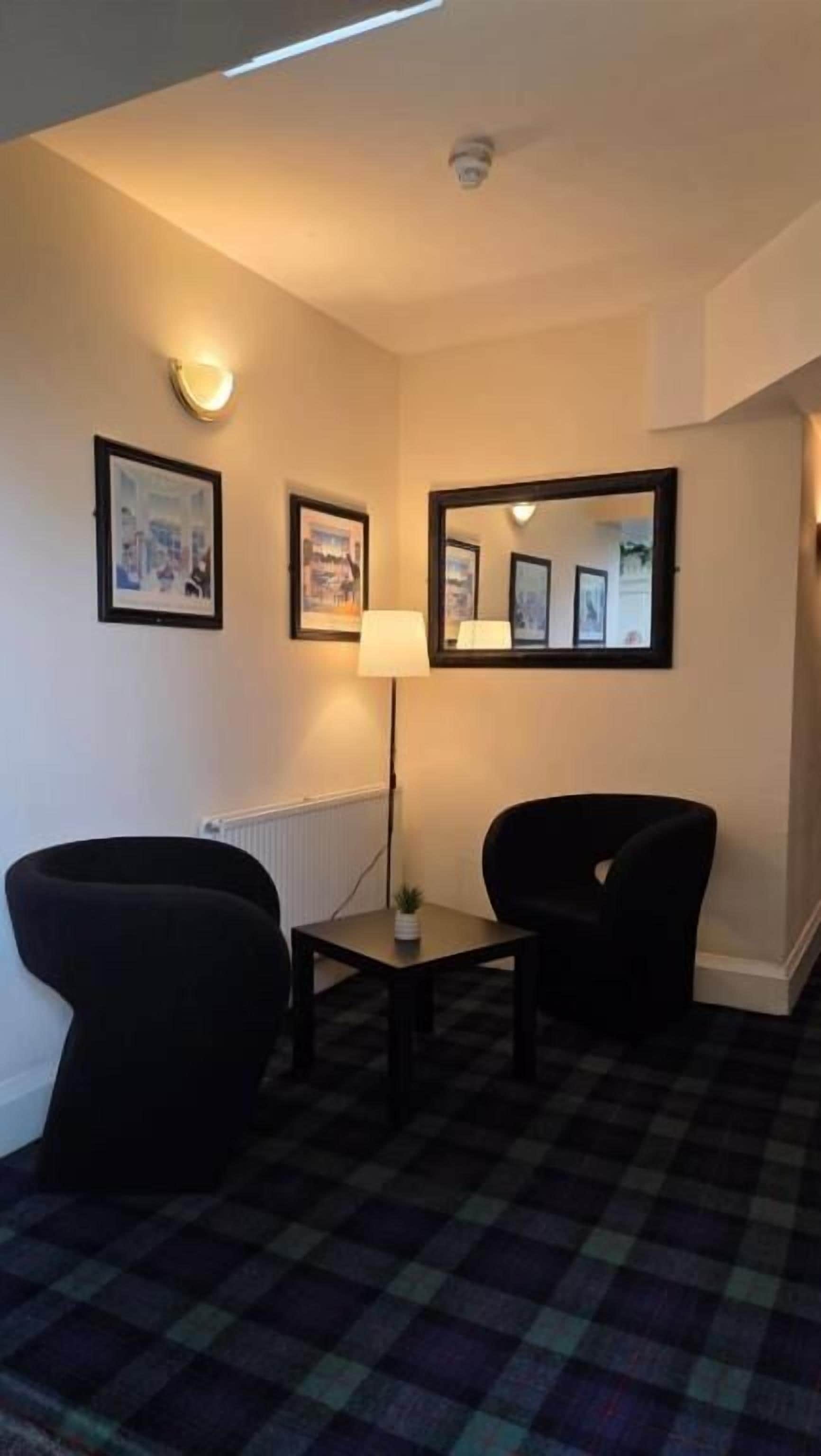 A hotel room with a checkered floor, two armchairs, a small table, a floor lamp, and framed pictures on the walls.