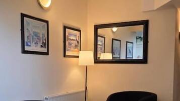 A hotel room with a checkered floor, two armchairs, a small table, a floor lamp, and framed pictures on the walls.