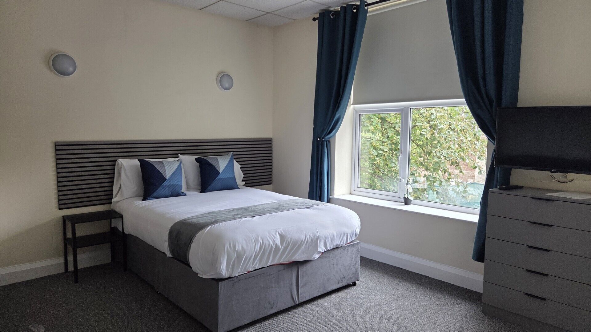 A bedroom with a bed, a dresser, a window with curtains, and a wall-mounted light fixture.