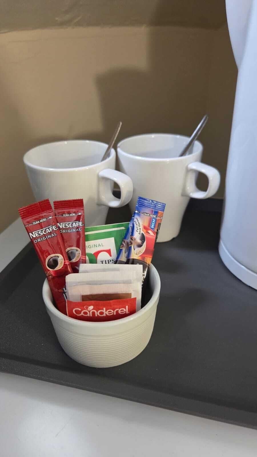 A tray with two white mugs, one with a spoon, and a cup containing coffee pods and a sachet.