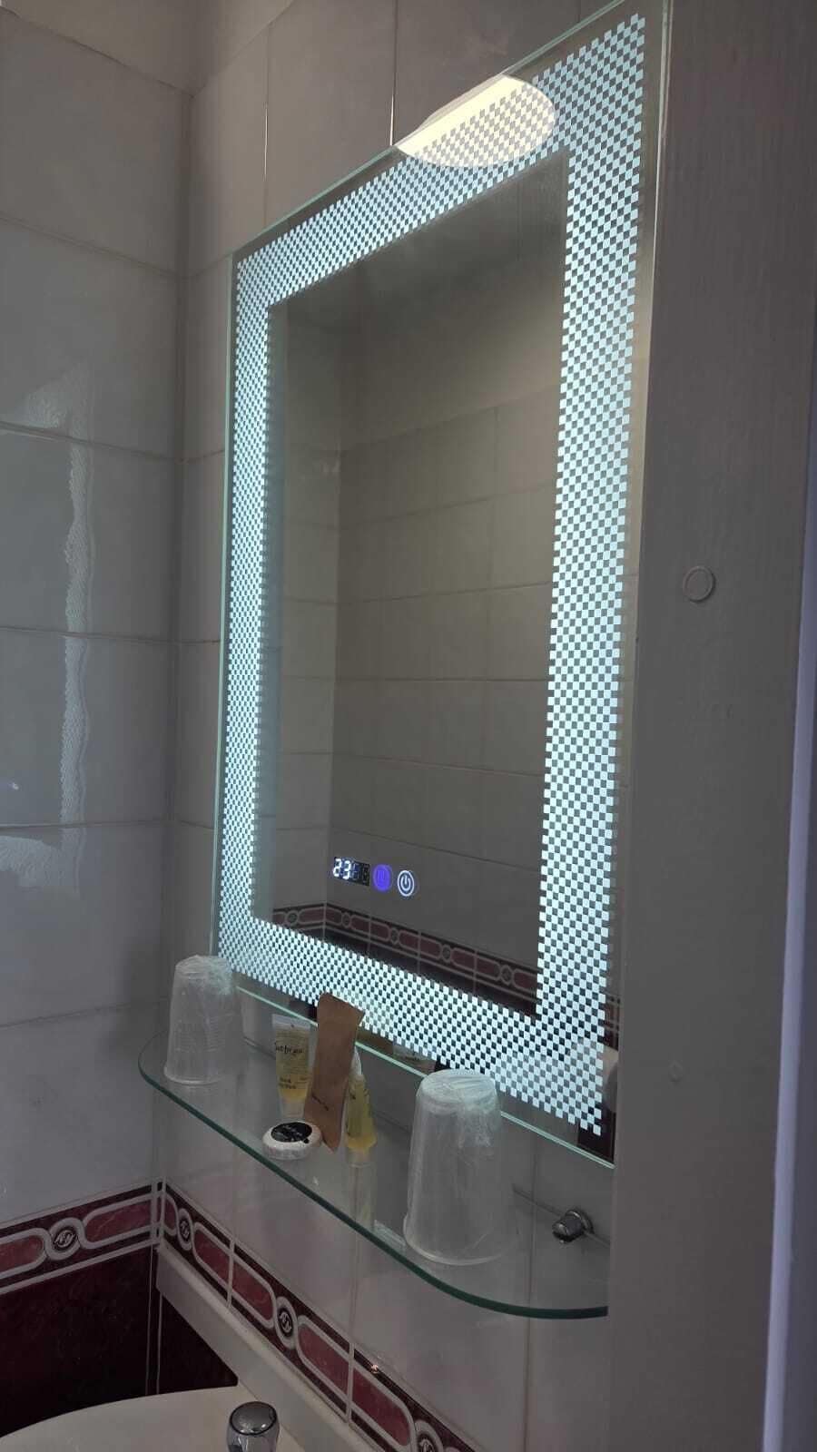 A bathroom with a large mirror, a glass shelf with toiletries, and a tiled wall.