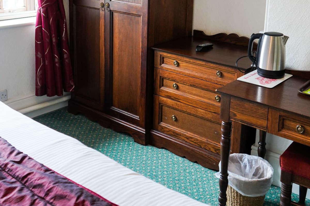 A bedroom with a wooden wardrobe, a television on the wall, a desk with a kettle, and a bed with a red and white duvet.