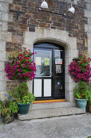 A stone building with a black door, two hanging flower baskets, and a small step leading to the entrance. - The Fox & Hounds Hotel (Okehampton)