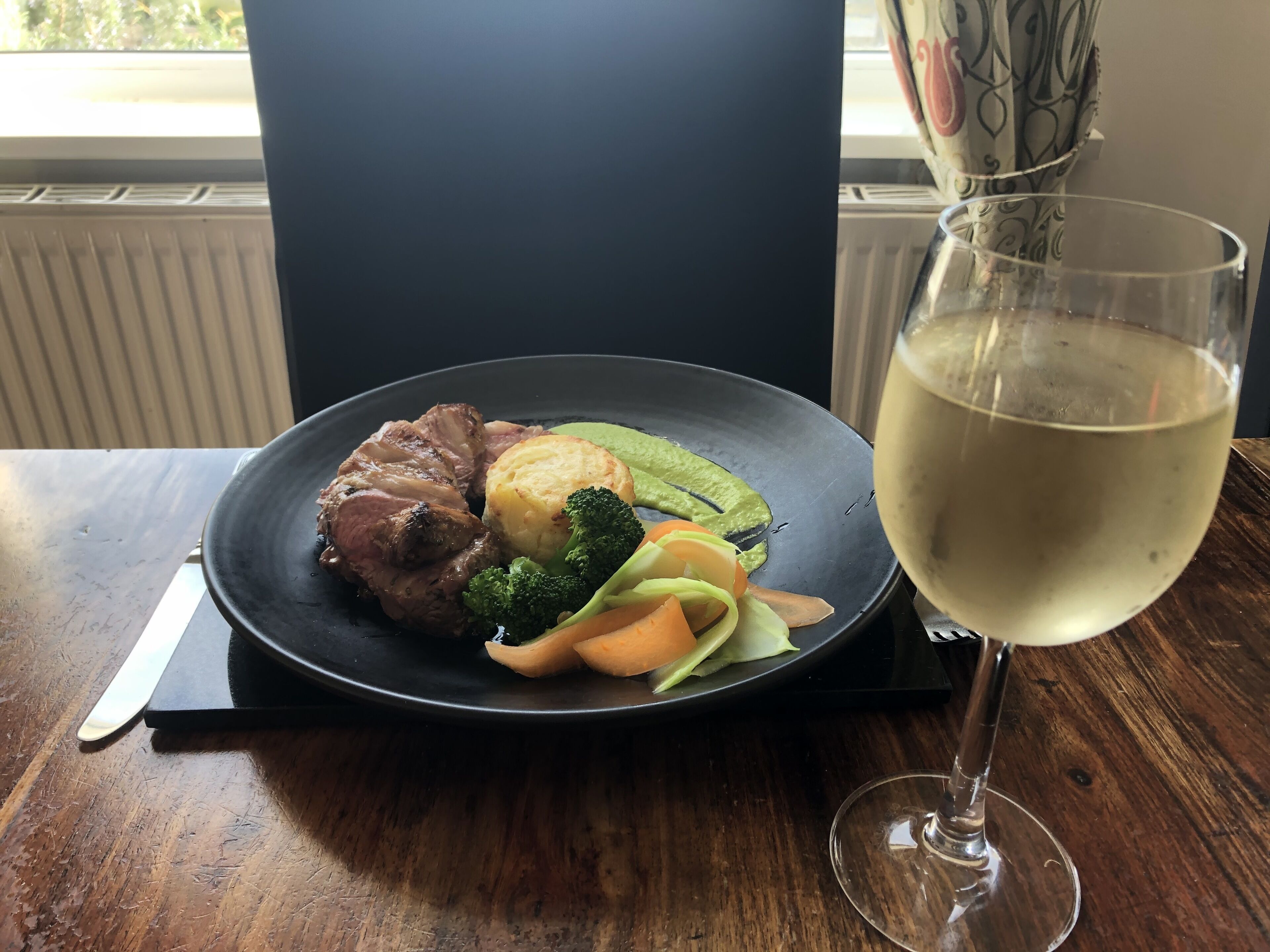 A plate of food with meat, vegetables, and a glass of white wine on a wooden table.