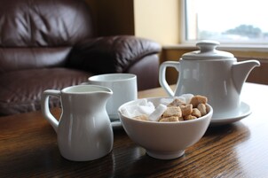 A wooden table with a white pitcher, a white cup, a white teapot, and a bowl of sugar cubes.