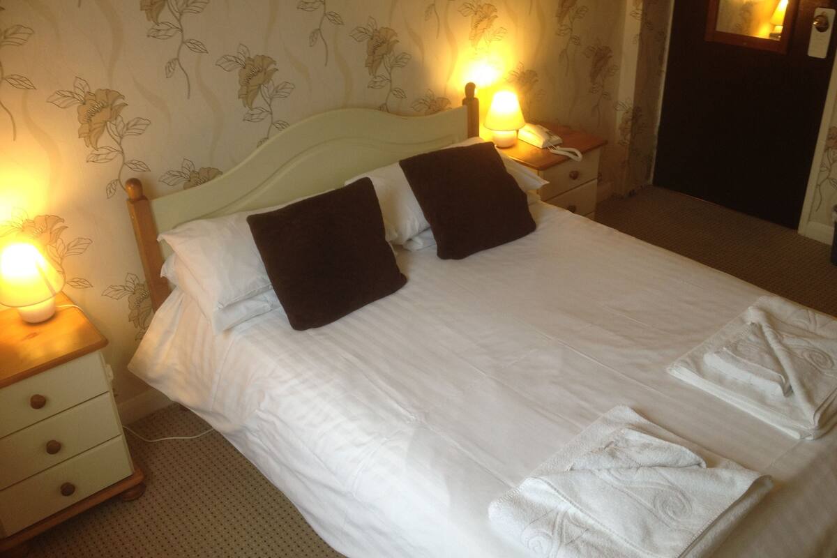 A neatly made bed with white linens and two dark brown pillows, flanked by bedside tables with lamps and a phone.